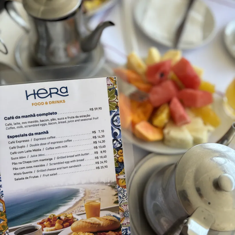 Cardápio de café da manhã do Hera sobre a mesa, com prato de frutas